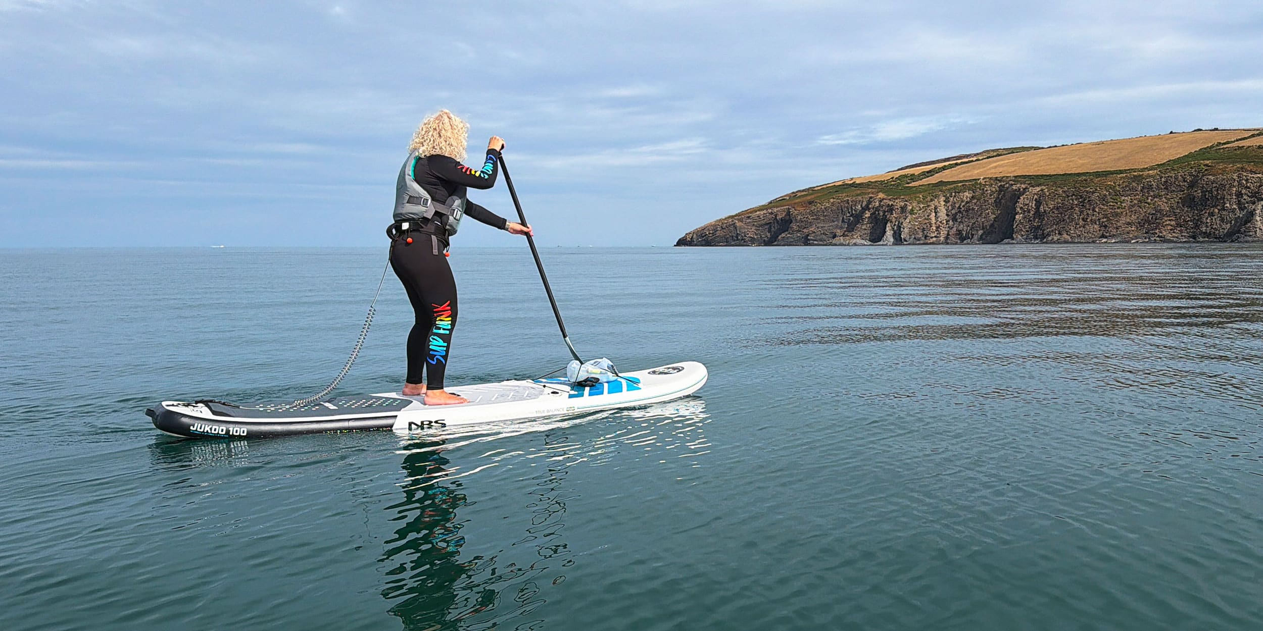 Paddleboarder on an inflatable paddle board in calm coastal water, with cliffs in the background.