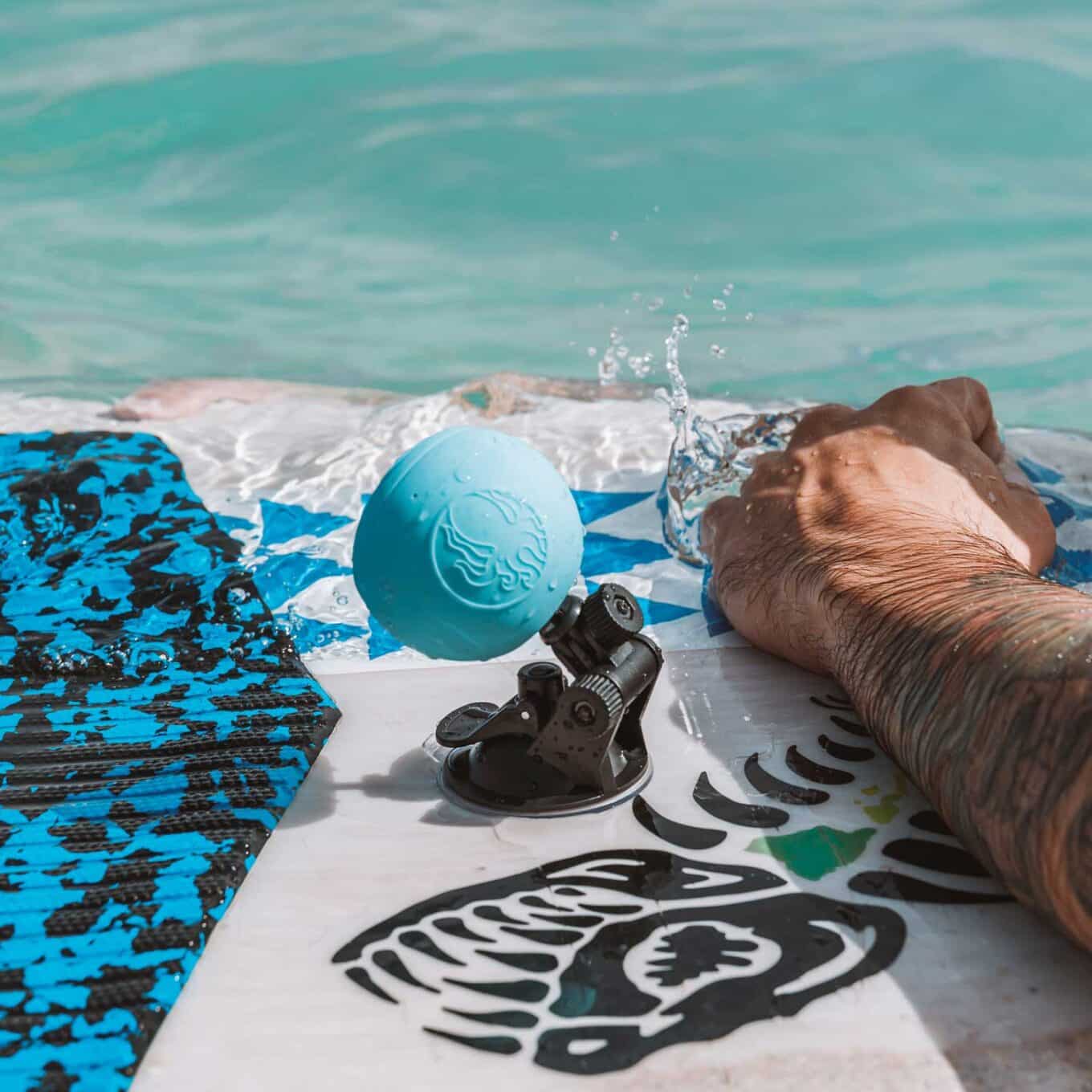 Blue speaker tilted on a Speaqua Board Mount as water washes over a paddle board beside a paddler’s arm